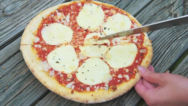 Female woman hand holding cutting with kitchen knife fresh crust whole small pizza on table with melted mozzarella cheese and sprinkles and red tomato sauce closeup as Italian food cuisine