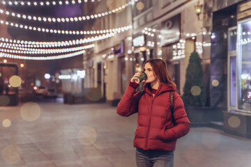 Fototapeta premium Girl in night Christmas city enjoy coffee and atmosphere. Young woman outdoors in winter. Lights on New Years Eve