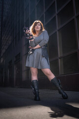 A curvy photographer girl in a gray short dress, leather platform boots and with a camera in her hands poses against the backdrop of an urban landscape with a skyscraper