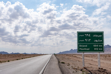 A road sign in Naein, Iran, captured in May 2018, with a clear sky and mountains in the background,...