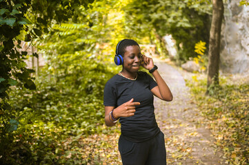 Attractive happy smiling young natural beauty short haired African Black woman with blue headphones in black t-shirt listening music and dancing in nature summer park