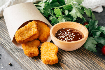 Serving of chicken nuggets in craft paper bag with barbecue sauce, sesame, cherry tomatoes, parsley on wooden surface