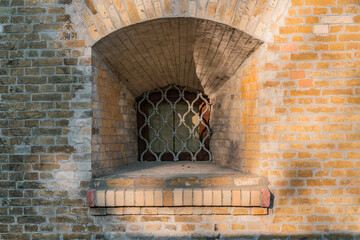 Wooden small arch window of medieval fortress with fat walls and decorative iron protection grate. Old. History. Architecture. Brick wall. Lattice of iron rods. Defence. Protection
