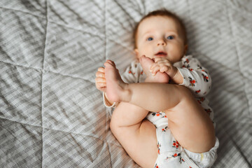 A little baby is lying on the bed in a patterned bodysuit and holding on to the legs. Tiny baby feet  close-up. The face is out of focus.