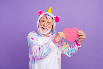 Portrait of attractive bearded cheerful grey-haired man in kigurumi holding heart amour isolated over violet purple color background