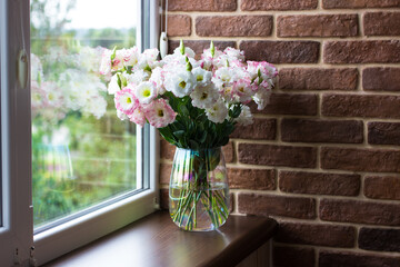 Eustomas in a vase on a brown windowsill by the window against the background of a brick wall
