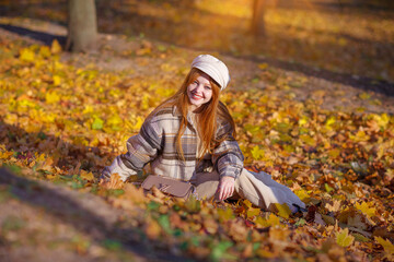 A beautifyl girl with long red hair walks in the park, around the yellow leaves of trees. The woman is dressed in a cap and a warm jacket in  cage. Joy, autumn, sunny day, vacation concept.