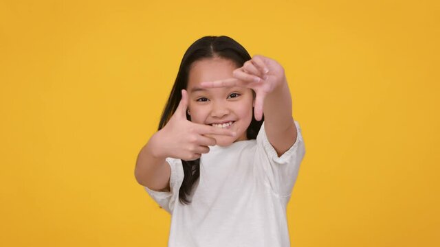 Smile to camera. Cute little asian girl making frame with fingers and smiling, pretending to make photography