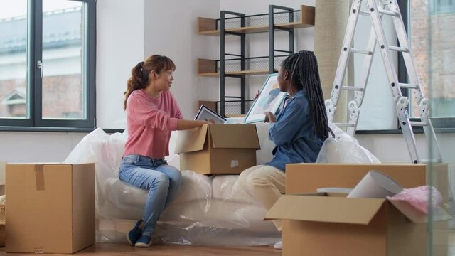 moving, people and real estate concept - women unpacking boxes with abstract pictures in frames at new home