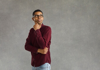 Studio portrait of pensive thoughtful young african american man in glasses touching chin looking over shoulder to side weighing pros and cons of sentence thinking. Advertisement concept