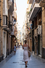 A lady in a white dress in a narrow, bustling street in Cádiz, Andalusia, filled with shops and pedestrians, showcasing the charming and vibrant city life