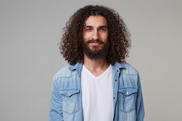 Indoor shot of young bearded male with curly long hair wears denim blue shirt smiles and looking directly into camera isolated over grey background