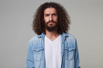 Indoor shot of young bearded male with curly long hair wears denim blue shirt looks sad with negative facial expression isolated over grey background