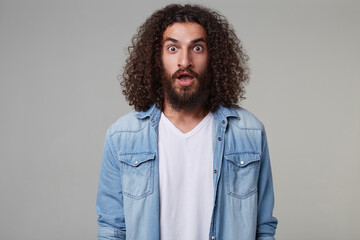 Fototapeta na wymiar Indoor shot of young bearded male with curly long hair wears denim blue shirt keeps his eyes and mouth widely opened with shocked facial expression. isolated over grey background