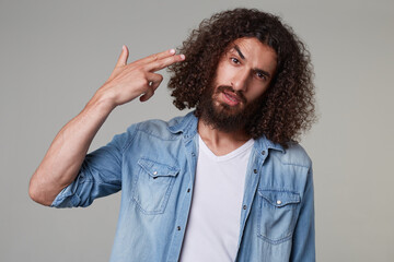 Fototapeta na wymiar Indoor shot of young bearded male with curly long hair wears denim blue shirt shows pistol sign and feels stressed and depressed. isolated over grey background