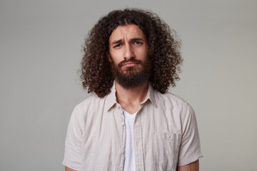 Indoor shot of young bearded male with curly long hair wears shirt with sad facial expression looks directly into camera. isolated over grey background