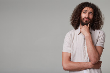 Indoor shot of young bearded male with curly long hair wears shirt looks aside at copy space, touching chin and think about future. isolated over grey background