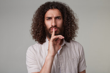 Indoor shot of young bearded male with curly long hair wears shirt negatively shows silence gesture. isolated over grey background