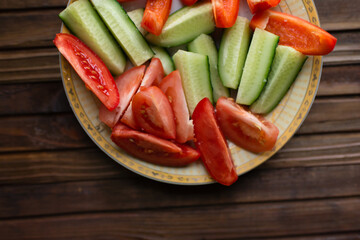 Vegetables on a plate. Tomato, pepper, cucumber cut into pieces. Sprinkled with salt. Vegetables on a plate on a brown wooden background. View from above.