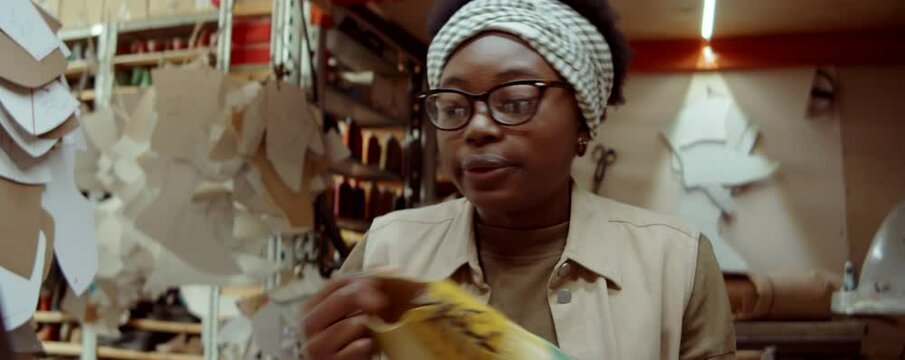 African American woman discussing new handmade shoes on online video call on laptop while working in shoemaker workshop