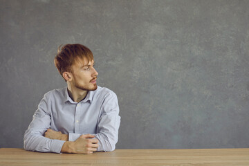 Seated casual young man serious looking to the side against gray background, copy space. For advertising and text. Bearded man looks to the side while sitting at the table.