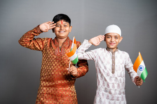 HIndu Muslim Children saluting by holding Indian flag on gray background - concept of kids celebrating Indian Republic or Independence day, Religious unity in diversity, oneness and patriotism.