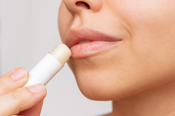 Cropped shot of a young caucasian beautiful woman applying a hygienic lipstick on her lips on a gray background. Moisturizing chapstick for dry lips - obrazy, fototapety, plakaty