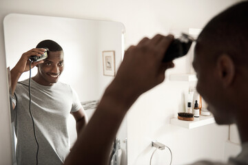 Smiling African man trimming his hair in his bathroom mirror - obrazy, fototapety, plakaty