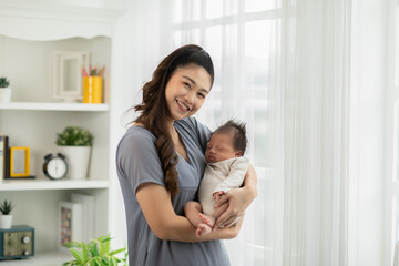 Obraz na płótnie Canvas Beautiful Asian woman holding newborn baby in her arms standing in front of windows at cozy home.Happy infant baby sleep in mother arms safety and comfortable.Mom and Baby Concept