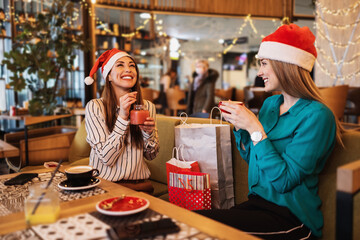 Two best friends with Santa hats sitting in coffee bar or restaurant after shopping and happily talking together. They also show each other the clothes they bought.