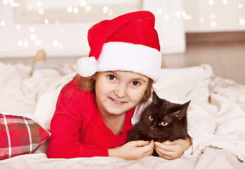 Little girl in funny Santa hat and Christmas pajamas sitting on a plaid with favorite pet black cat