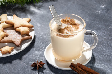 Glass mug with traditional Christmas drink eggnog with cinnamon and heart shape cookies on grey.