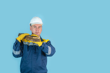 A carpenter in a hardhat and gloves holds a stack of painted boards on his shoulder, looking into the camera. Copyspace on a blue background