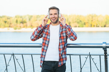 happy young trendy male fashion model in music headphones and casual shirt outside, music