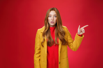 Impressed and questioned amazed redhead woman remain speechless from amazement pointing right open mouth in stupor, asking question about confusing shocking promotion over red wall