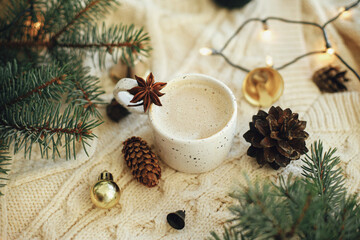 Obraz na płótnie Canvas Warm coffee in stylish cup with anise star, fir branches, ornaments, pine cones and warm lights on cozy knitted background. Atmospheric christmas time and hygge winter home