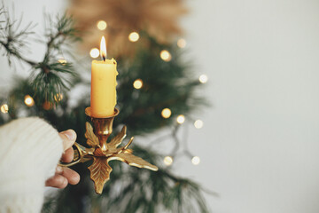 Atmospheric hygge winter home. Hand in cozy sweater holding vintage candlestick with burning candle on background of warm lights, fir branches, sweden star in festive scandinavian room