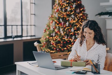 Pleased european woman office worker writing christmas letter, greeting card with holiday wishes