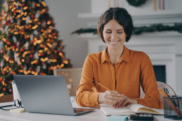 Smiling italian female making notes, using laptop at home office during christmas holidays