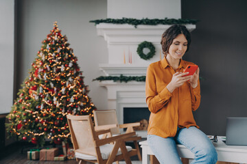 Smiling hispanic woman using mobile phone during remote work at home on Christmas holidays