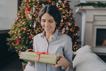 Smiling european woman sitting on sofa in room decorated for xmas holidays holding Christmas present