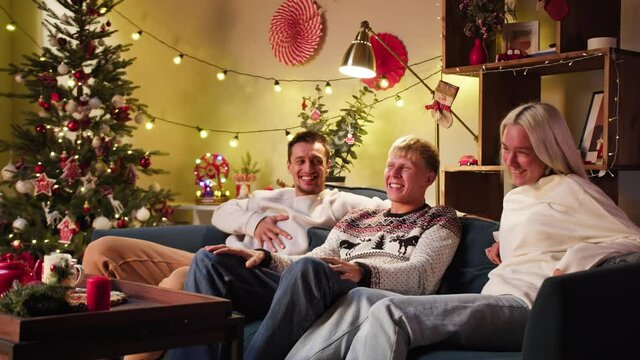 Friends watching television, comedy film. Young people sitting on sofa, watch christmas movie on tv, laughing. Men and women during New Year time at home. Cozy winter evening concept, holidays. 