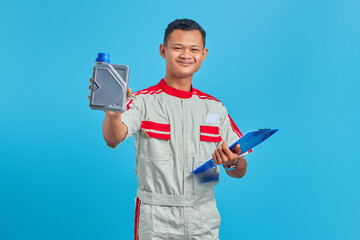 Portrait of smilling young asian mechanic holding clipboard and showing plastic bottle of engine oil in hand over blue background