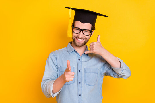 Photo of young man happy smile show fingers call you sign wear hat master degree isolated over yellow color background