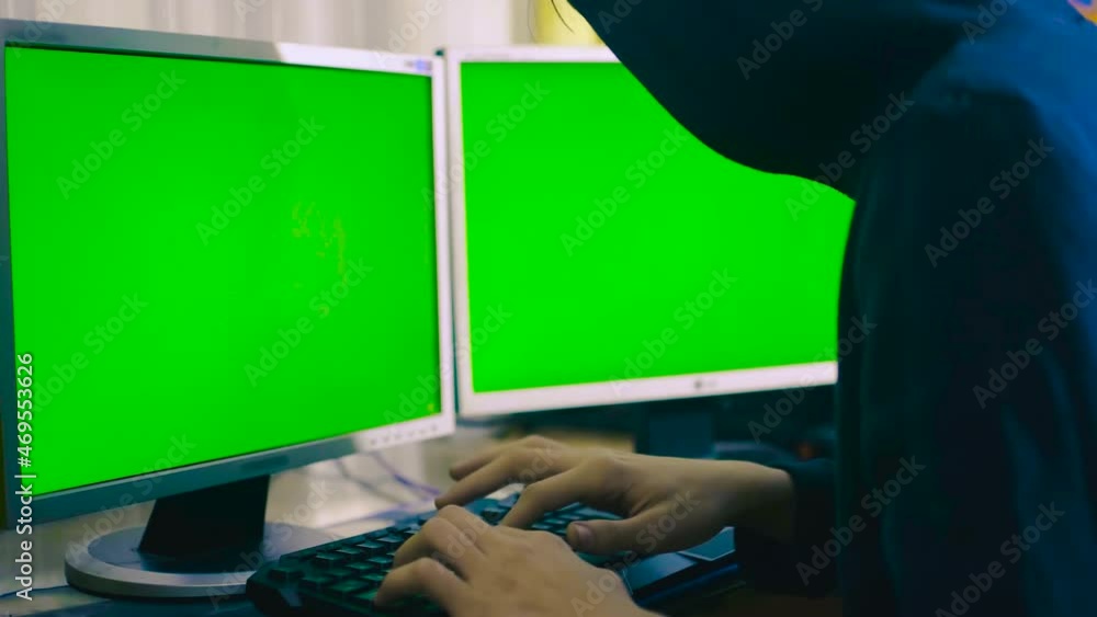 Poster a man is sitting at a table in a hood and typing on a keyboard, there are two monitors on the table