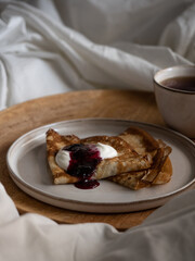 Breakfast in bed, a cup of black tea and thin pancakes with currant jam and sour cream on a beige ceramic plate on a wooden tray on a soft white bed in the morning light