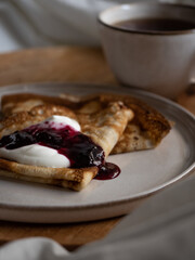 Breakfast in bed, a cup of black tea and thin pancakes with currant jam and sour cream on a beige ceramic plate on a wooden tray on a soft white bed in the morning light