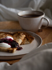 Breakfast in bed, a cup of black tea and thin pancakes with currant jam and sour cream on a beige ceramic plate on a wooden tray on a soft white bed in the morning light
