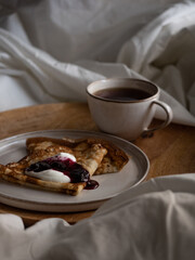 Breakfast in bed, a cup of black tea and thin pancakes with currant jam and sour cream on a beige ceramic plate on a wooden tray on a soft white bed in the morning light