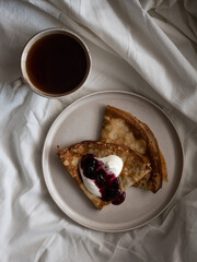 Breakfast in bed, a cup of black tea and thin pancakes with currant jam and sour cream on a beige ceramic plate on a soft white bed in the morning light